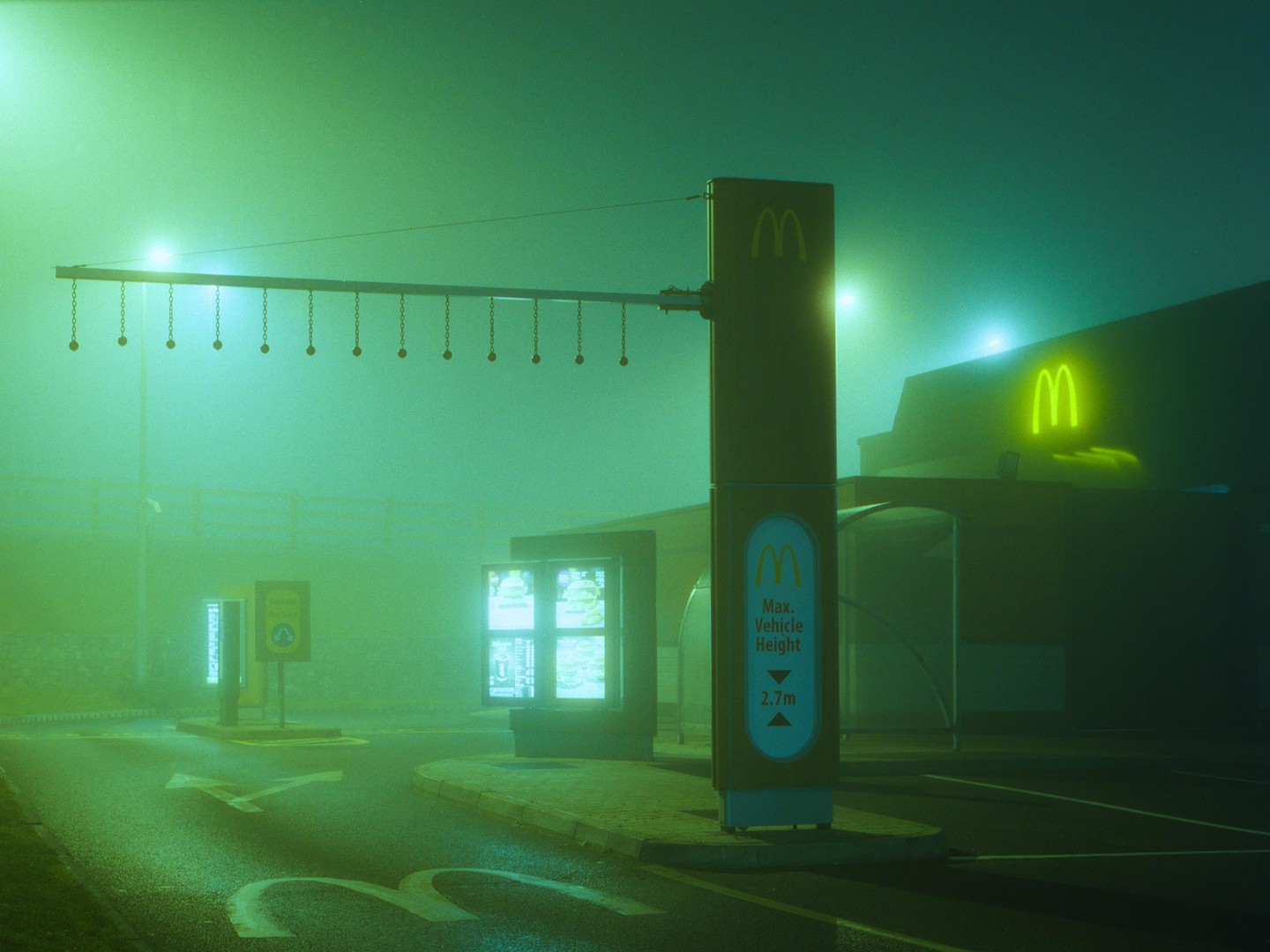 A McDonald's drive-thru is illuminated by green light and neon signs on a foggy night.