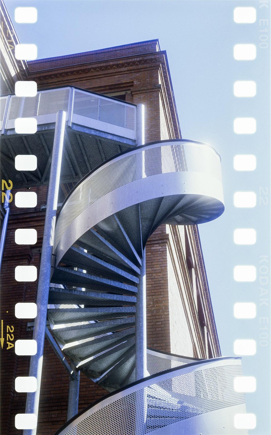 A metal spiral staircase with perforated railings ascends alongside a brick building under a clear sky.