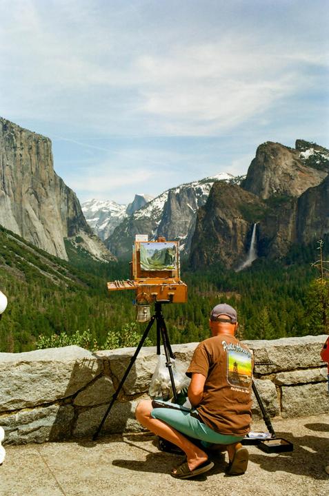 A man paints a landscape of a valley with mountains, trees, and a waterfall.