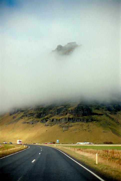 A winding road leads towards a large, fog-covered mountain with green and brown slopes.