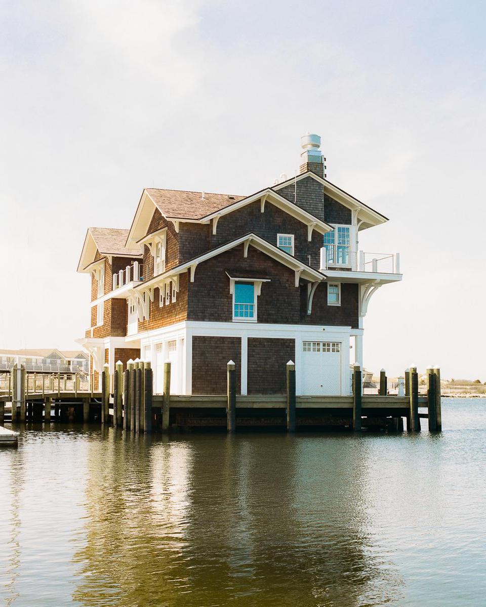 A large house with a brown shingle exterior and white trim stands on stilts over calm water, with a dock extending from its side.