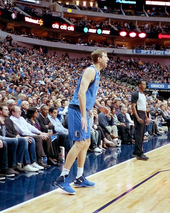 A basketball player in a blue uniform stands on the court during a game, with spectators in the background.