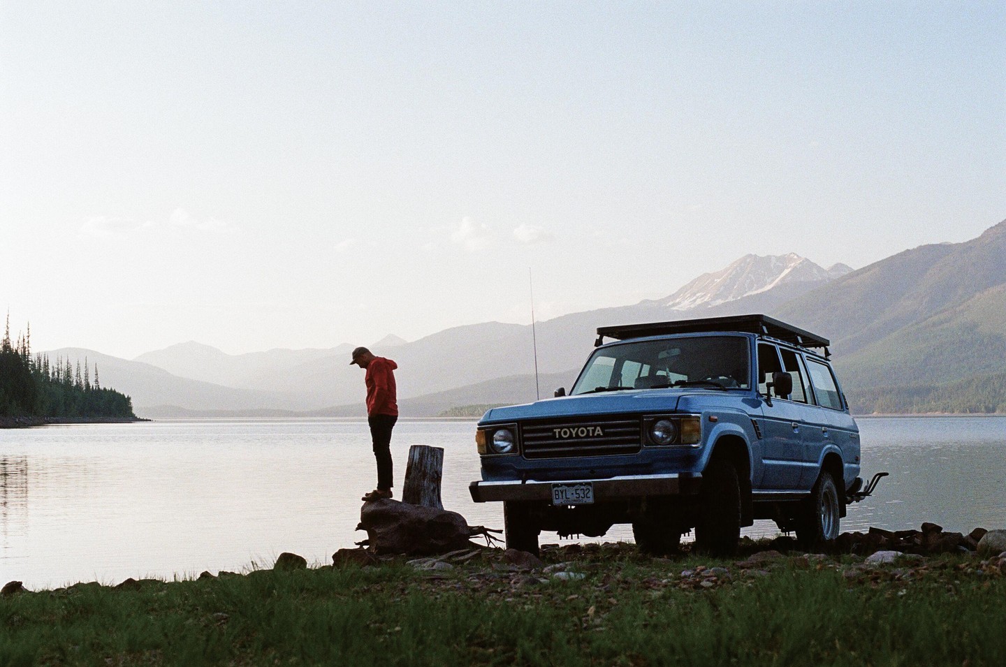 A man stands on a tree stump by a lake next to a blue Toyota SUV with mountains in the background.
