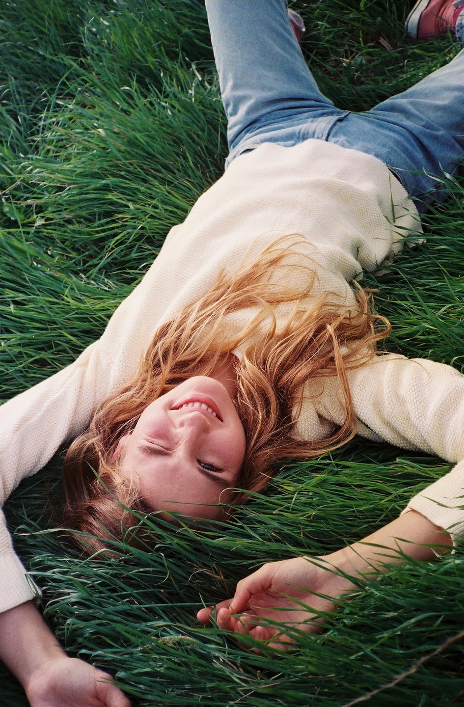 A young woman with long hair lies on her back in green grass, smiling at the camera.