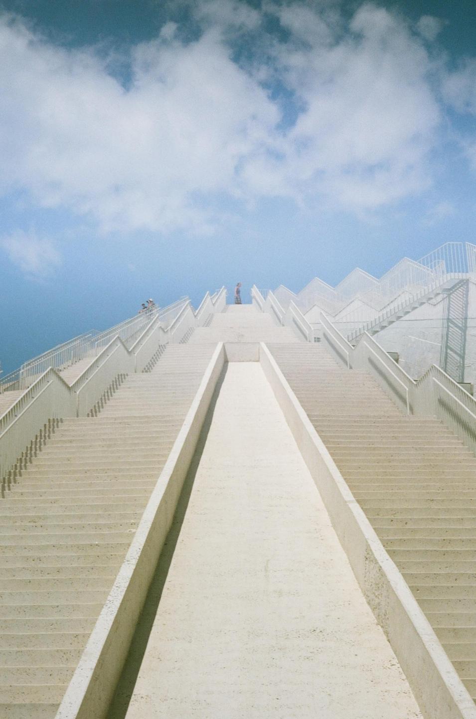 A person stands at the top of a wide, white, zig-zagging staircase under a blue sky with clouds.