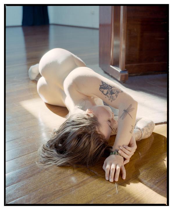 A woman with tattoos is lying on a wooden floor in a sunlit room.