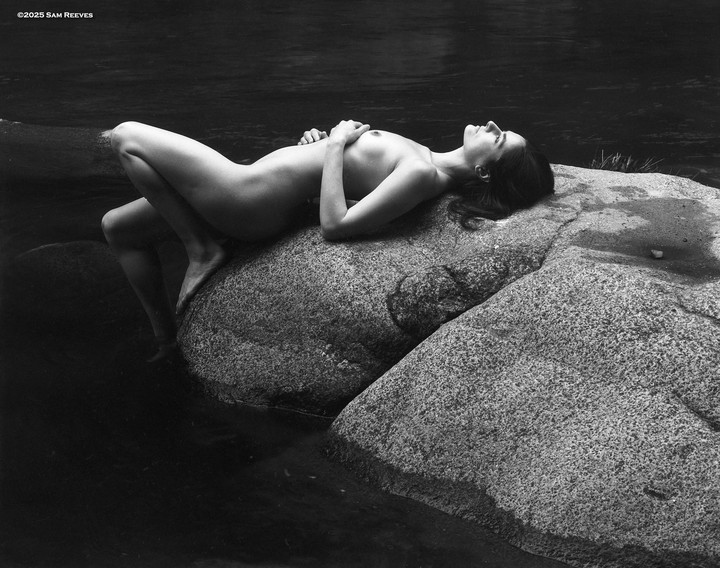 A nude woman lies on a large rock with her feet in the water of the San Lorenzo River.