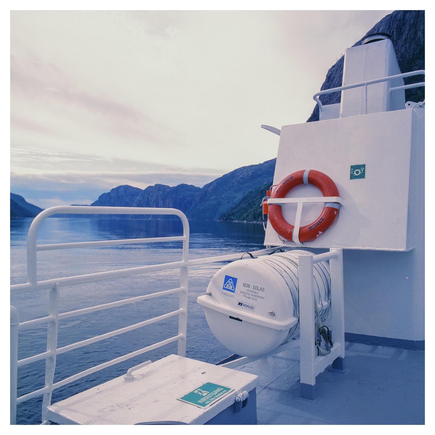 A ferry deck with a life preserver and a life raft overlooks a fjord surrounded by mountains.
