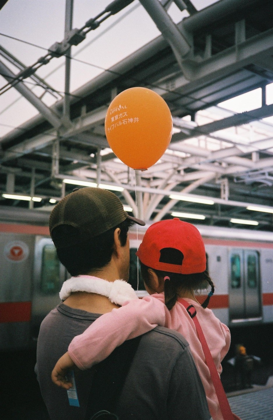 A man holds a girl on his shoulders while standing on a train platform with an orange balloon.