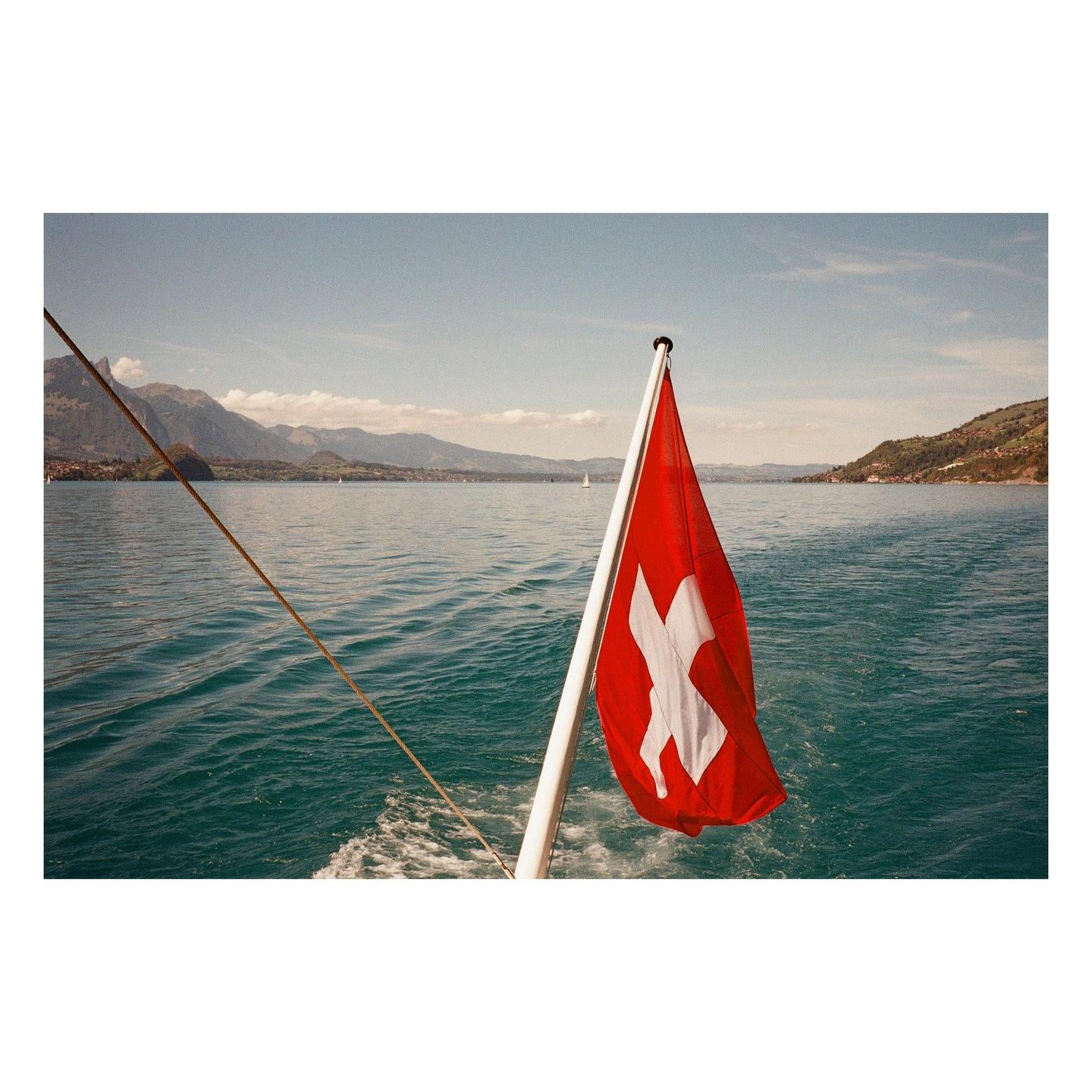 A Swiss flag flies from the back of a boat on a lake with mountains in the distance.