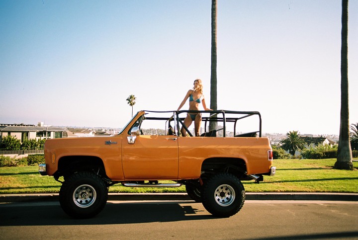 A woman in a bikini stands in the back of an orange pickup truck on a sunny day with palm trees in the background.