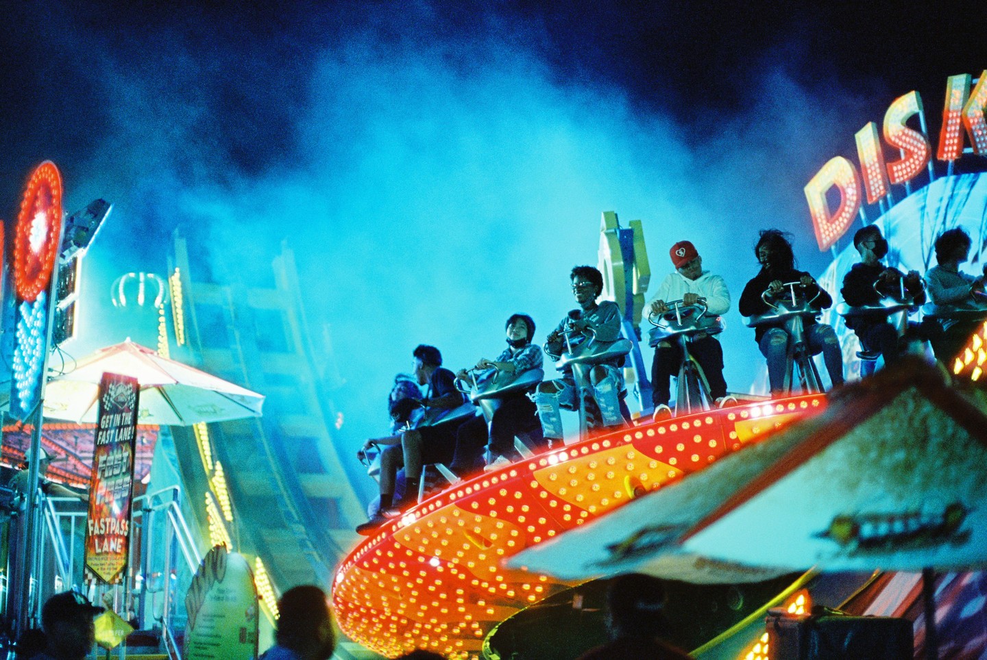 People ride a spinning carnival ride at night, illuminated by bright lights and a hazy blue sky.