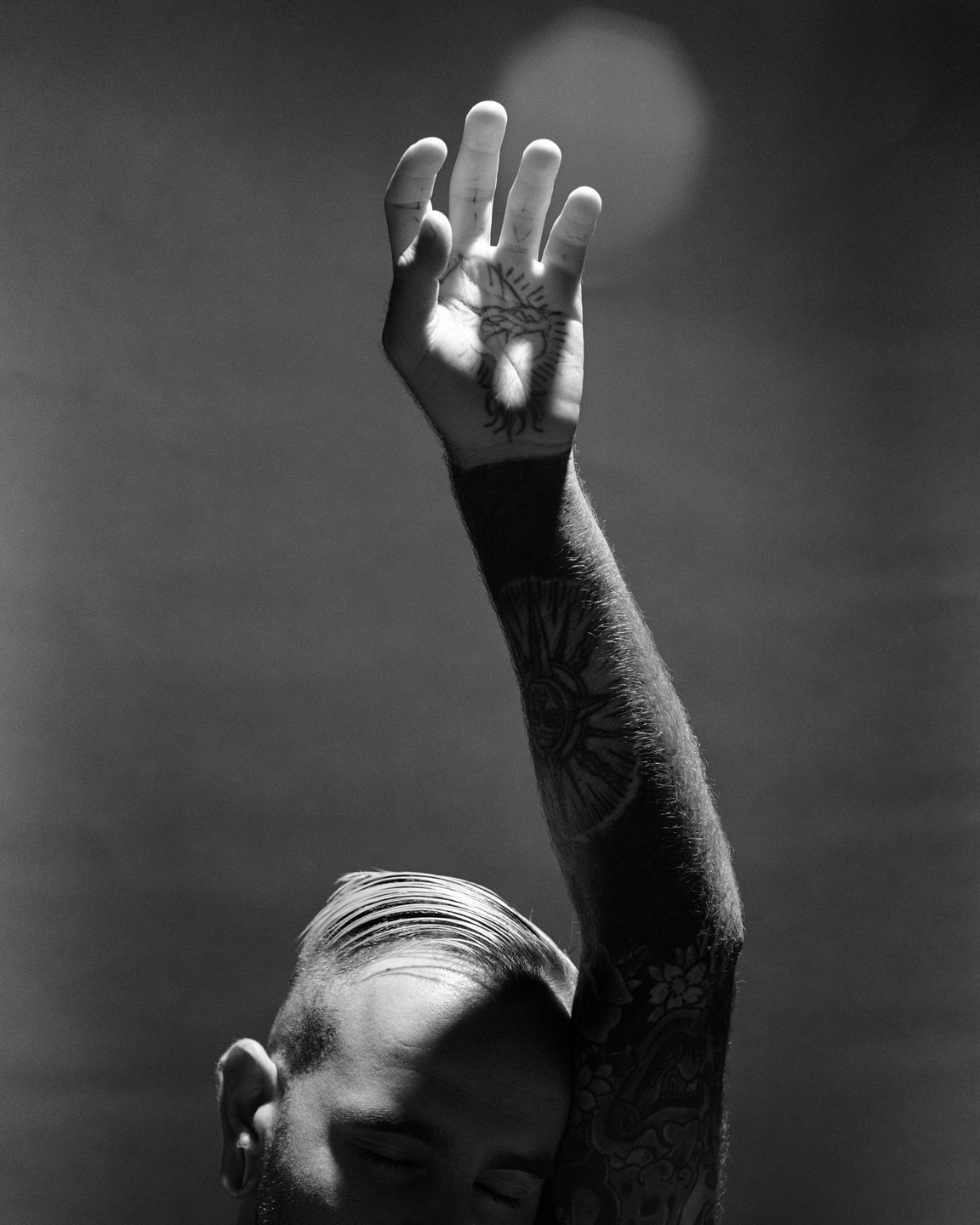 A man with tattoos on his arm and hand raises his arm, with his eyes closed and his head tilted back.