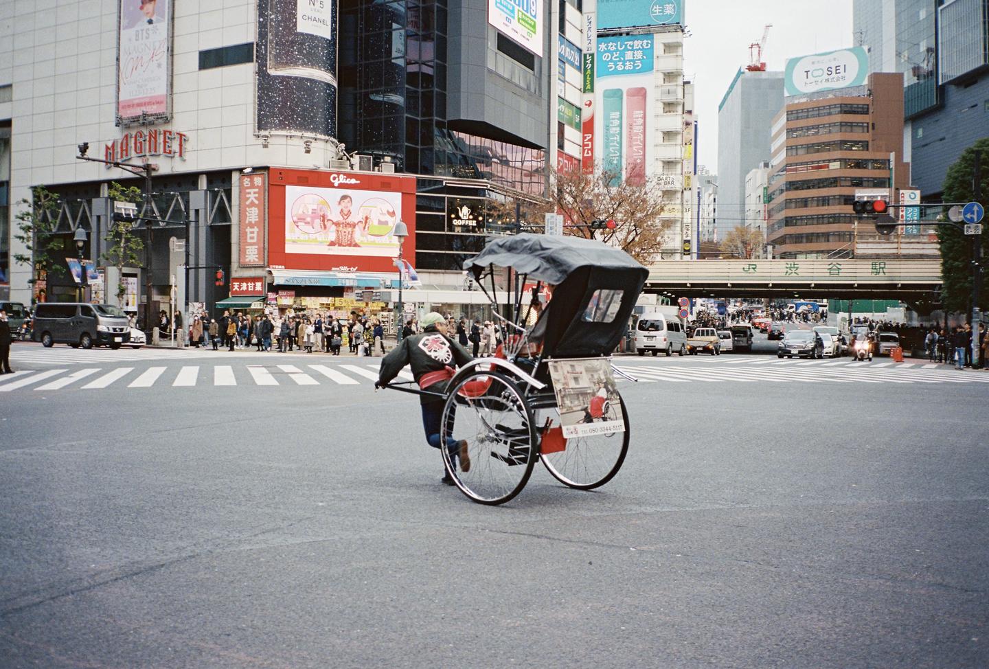 A man pulls a rickshaw across a street in a city with tall buildings and many people.