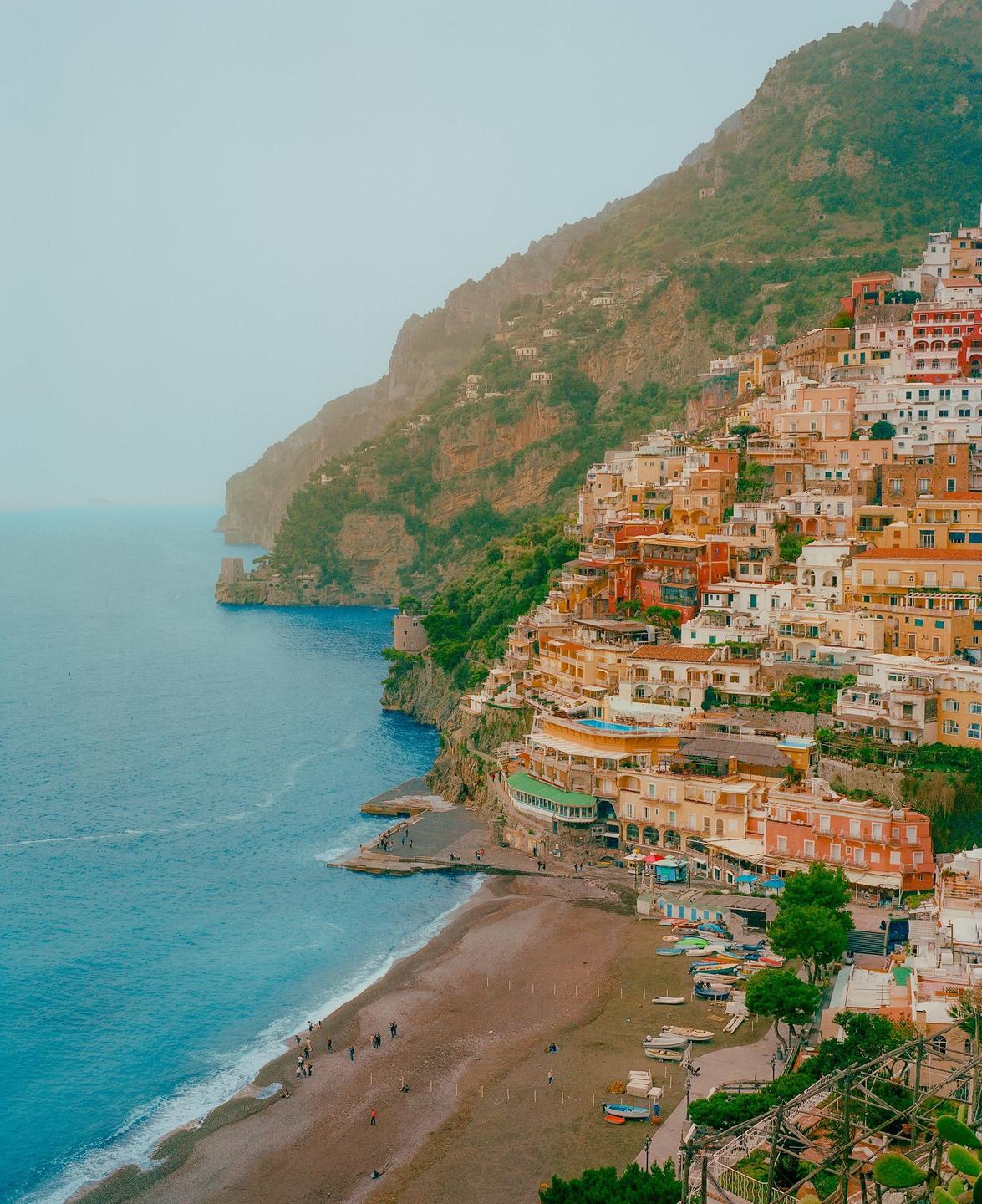A coastal town with colorful buildings is nestled into a steep hillside overlooking a beach and the sea.
