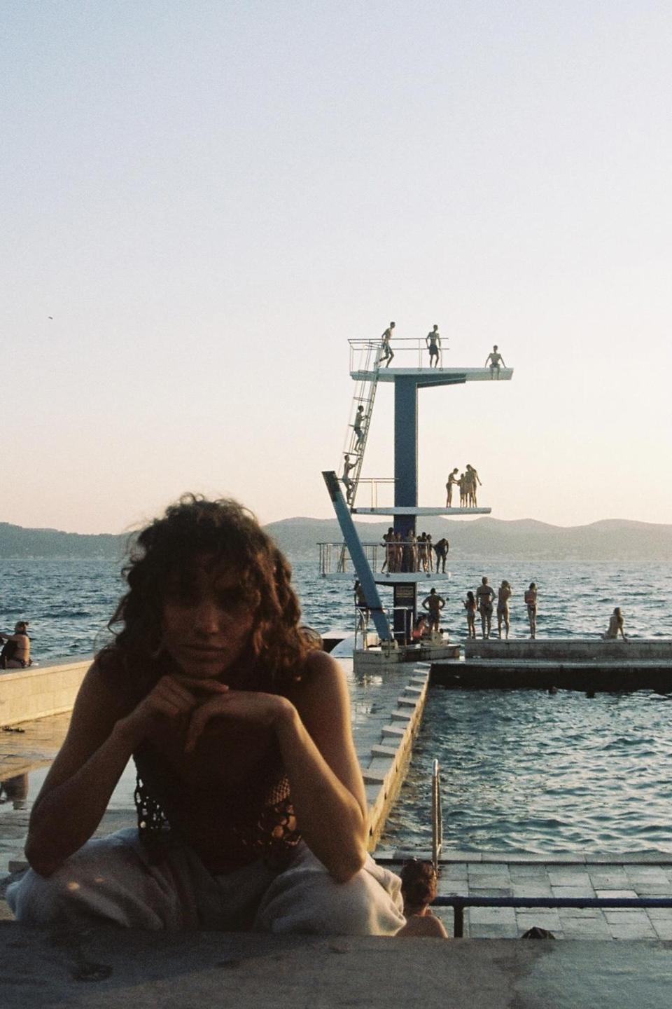 A woman sits in the foreground with a diving platform and people in the water behind her.