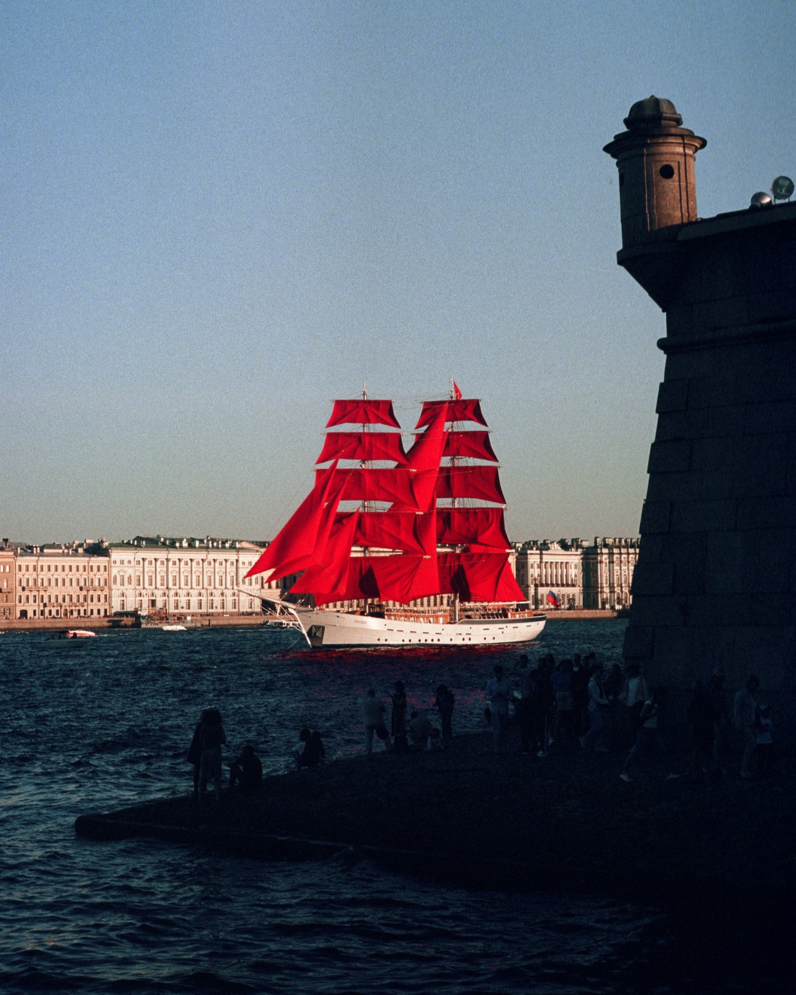 A large white sailboat with bright red sails moves across the water past a stone fortress and a city skyline.