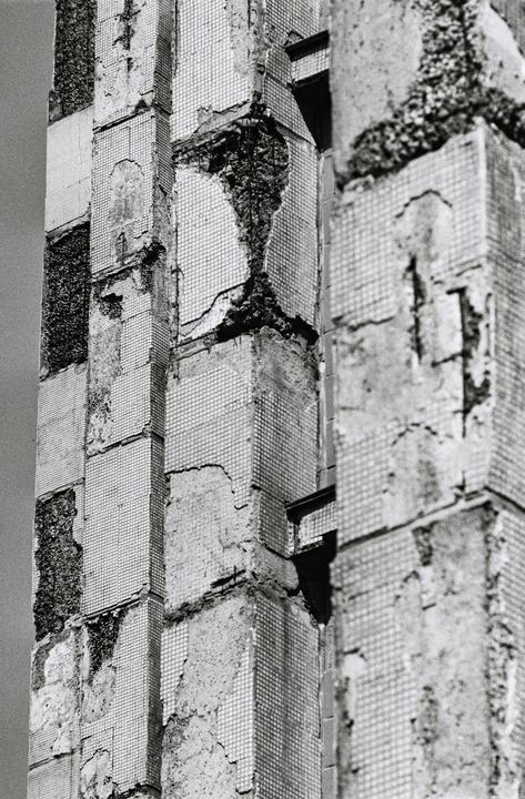 A close-up view shows the decaying facade of a building covered in small square tiles, with large sections of the tiles missing and the underlying concrete exposed.