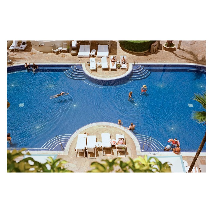A high-angle view shows people swimming and relaxing by a blue swimming pool with lounge chairs.