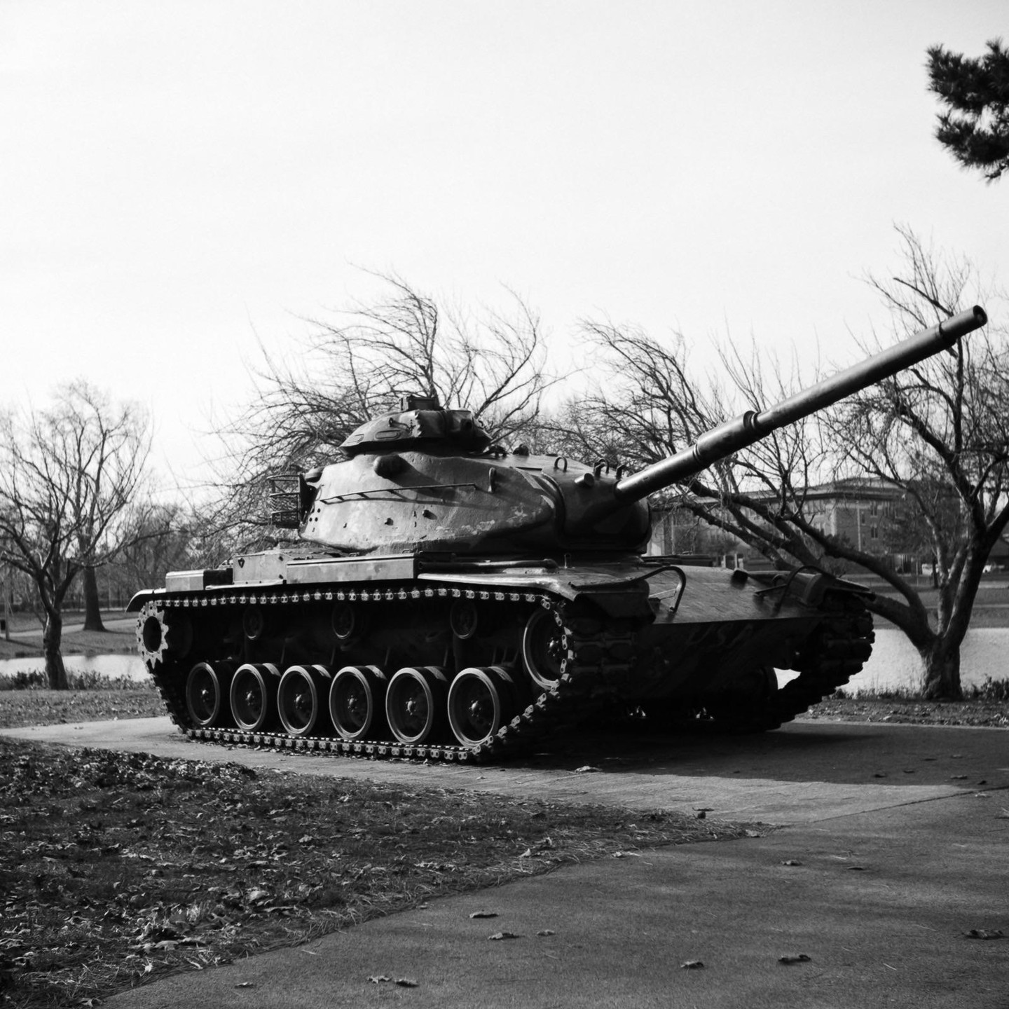 A tank is parked on a paved path next to a grassy area with bare trees and a body of water in the background.