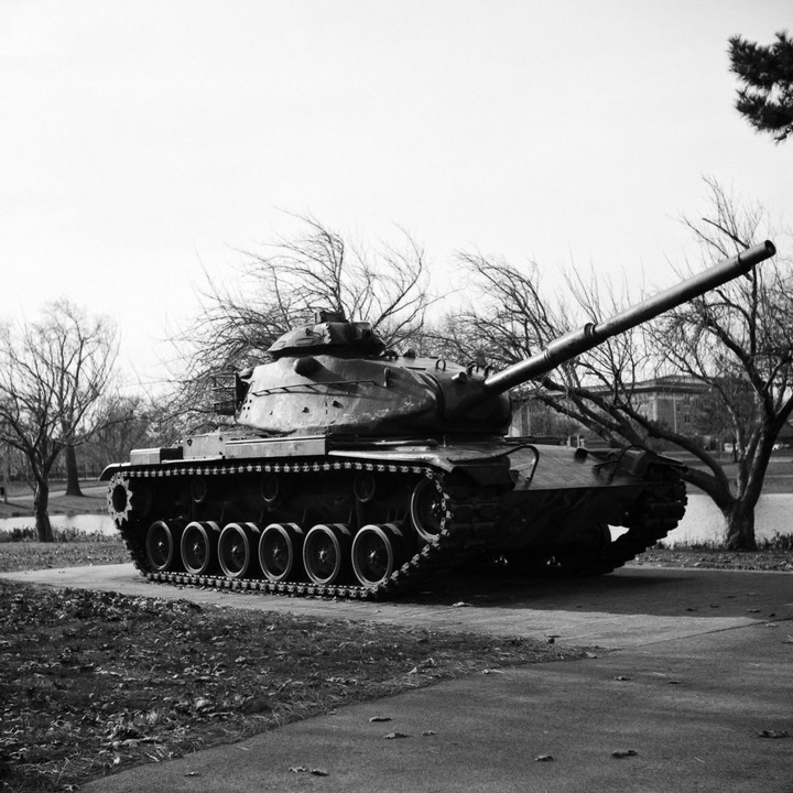 A tank is parked on a paved path next to a grassy area with bare trees and a body of water in the background.