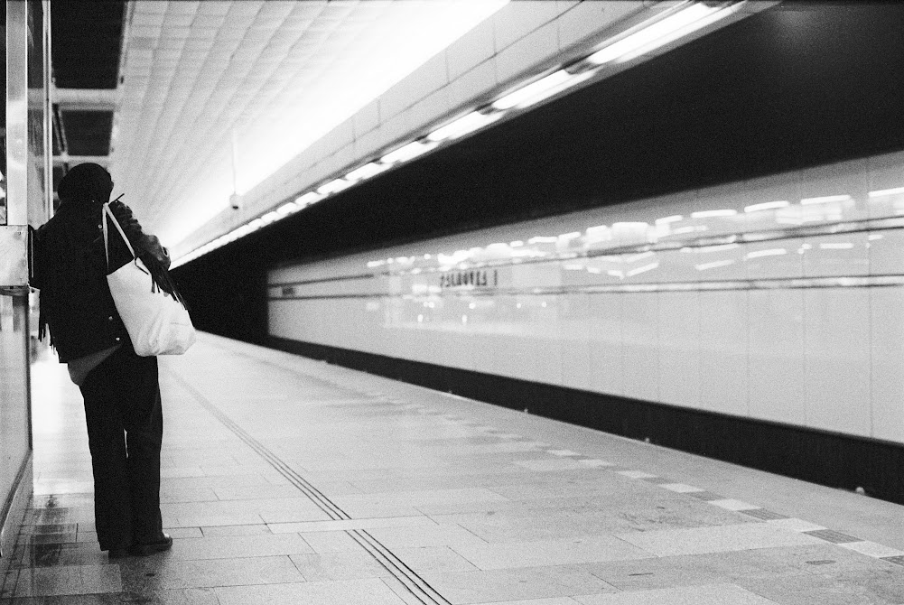 A person stands on a subway platform with a white bag over their shoulder, facing away from the camera.