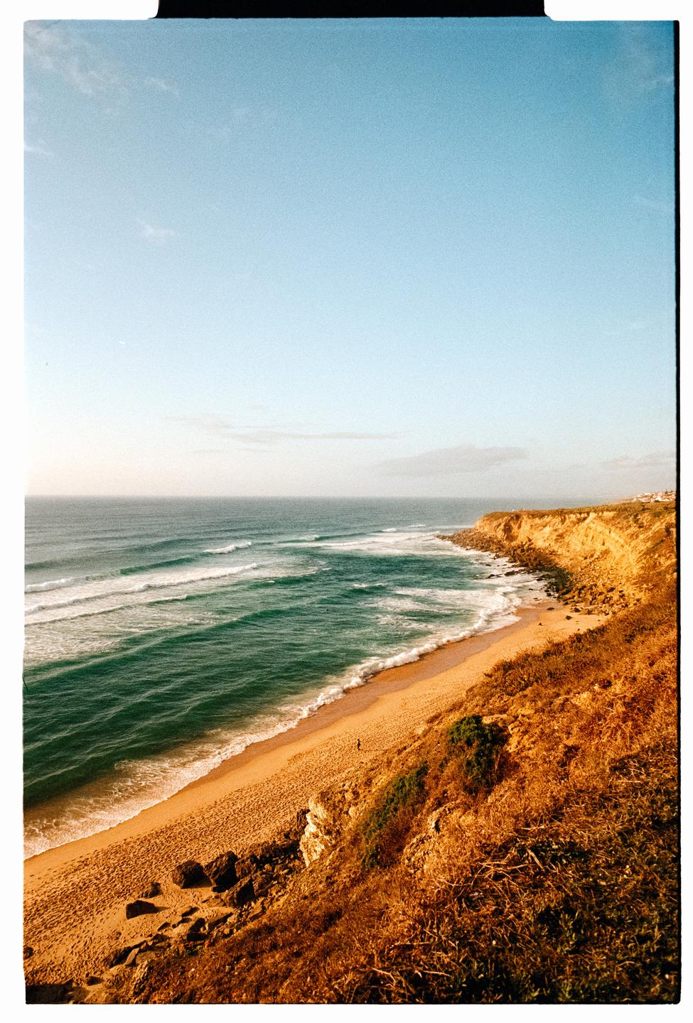 A sandy beach with waves crashing on the shore is visible from a high vantage point, with a cliffside covered in dry grass to the right.