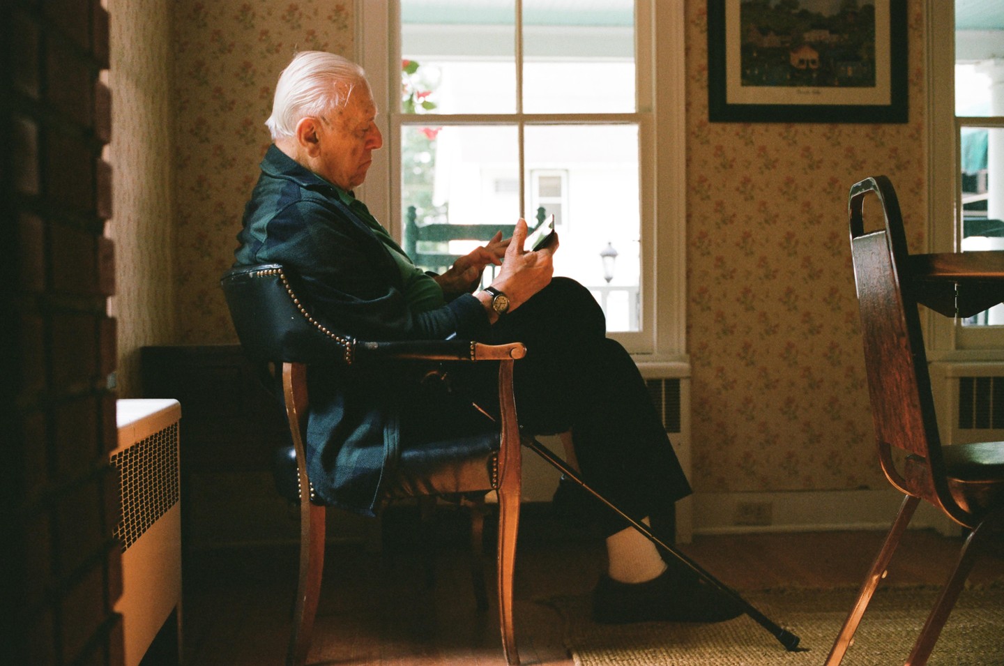 An elderly man sits in a chair by a window, looking at his phone.