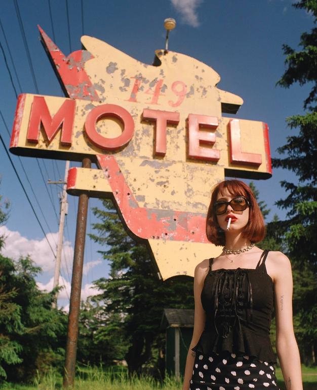 A woman with a cigarette in her mouth stands in front of a weathered motel sign.