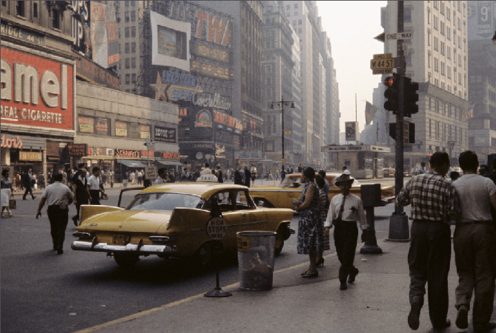 A busy street scene in New York City shows pedestrians, yellow taxis, and numerous billboards and signs on tall buildings.
