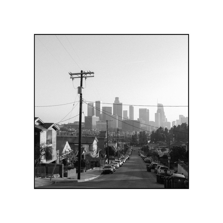 A street with parked cars leads down a hill towards a distant city skyline.