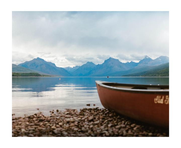 A red canoe rests on a rocky shore with a lake and mountains in the background under a cloudy sky.