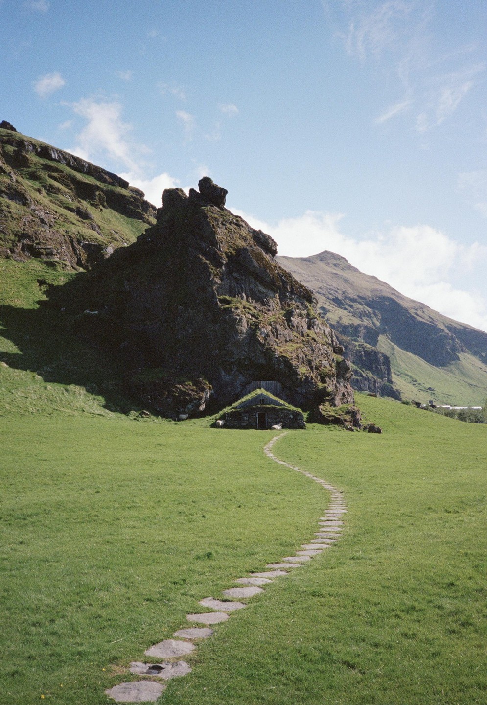 A stone path leads to a small cottage with a grass roof nestled at the base of a large rock formation in a green field.