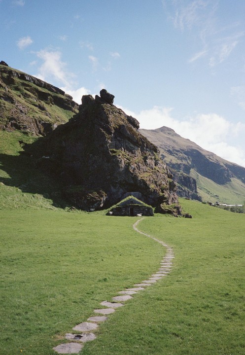 A stone path leads to a small cottage with a grass roof nestled at the base of a large rock formation in a green field.