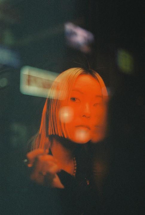 A woman with blonde hair looks directly at the viewer, with orange light reflecting on her face and bokeh in the background.