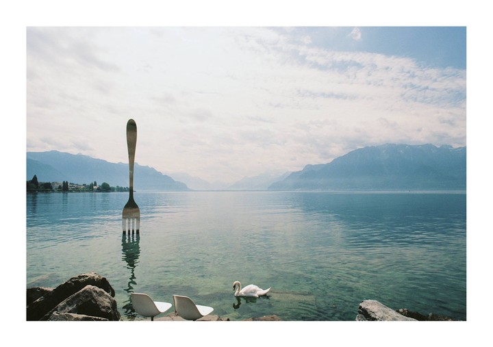 A giant fork sculpture stands in a lake with a swan swimming nearby and mountains in the background.