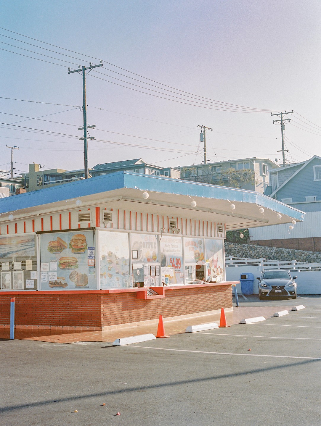 A drive-through restaurant with a blue roof and red brick counter stands in a parking lot with a car and orange cones.