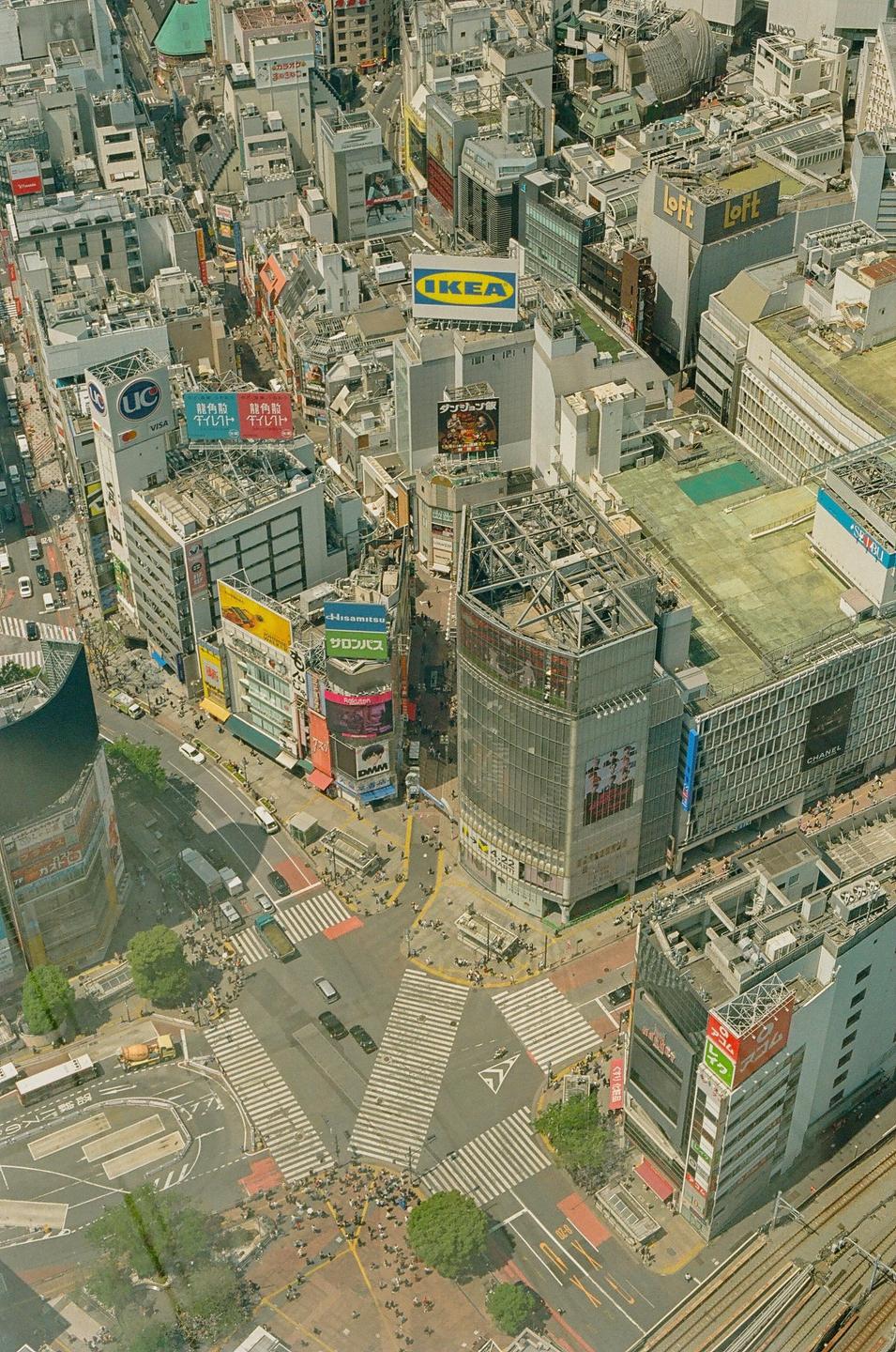 An aerial view shows a busy intersection with many pedestrians crossing the street and numerous buildings with advertisements.