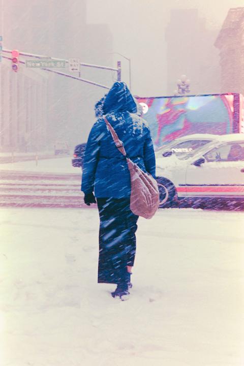 A person in a blue coat and black pants walks through a snowy street with cars and buildings in the background.