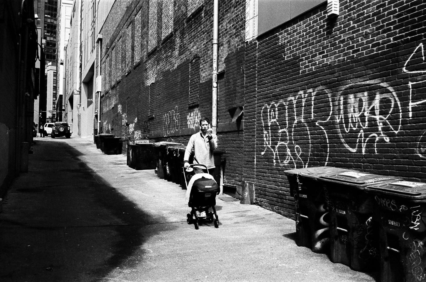 A man pushes a stroller down a narrow alleyway lined with brick buildings and graffiti-covered dumpsters.