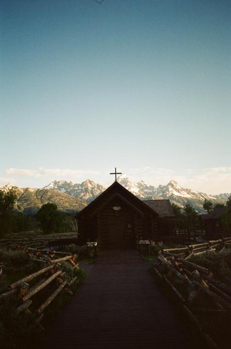 A wooden chapel with a cross on its roof stands before a mountain range with snow-capped peaks.