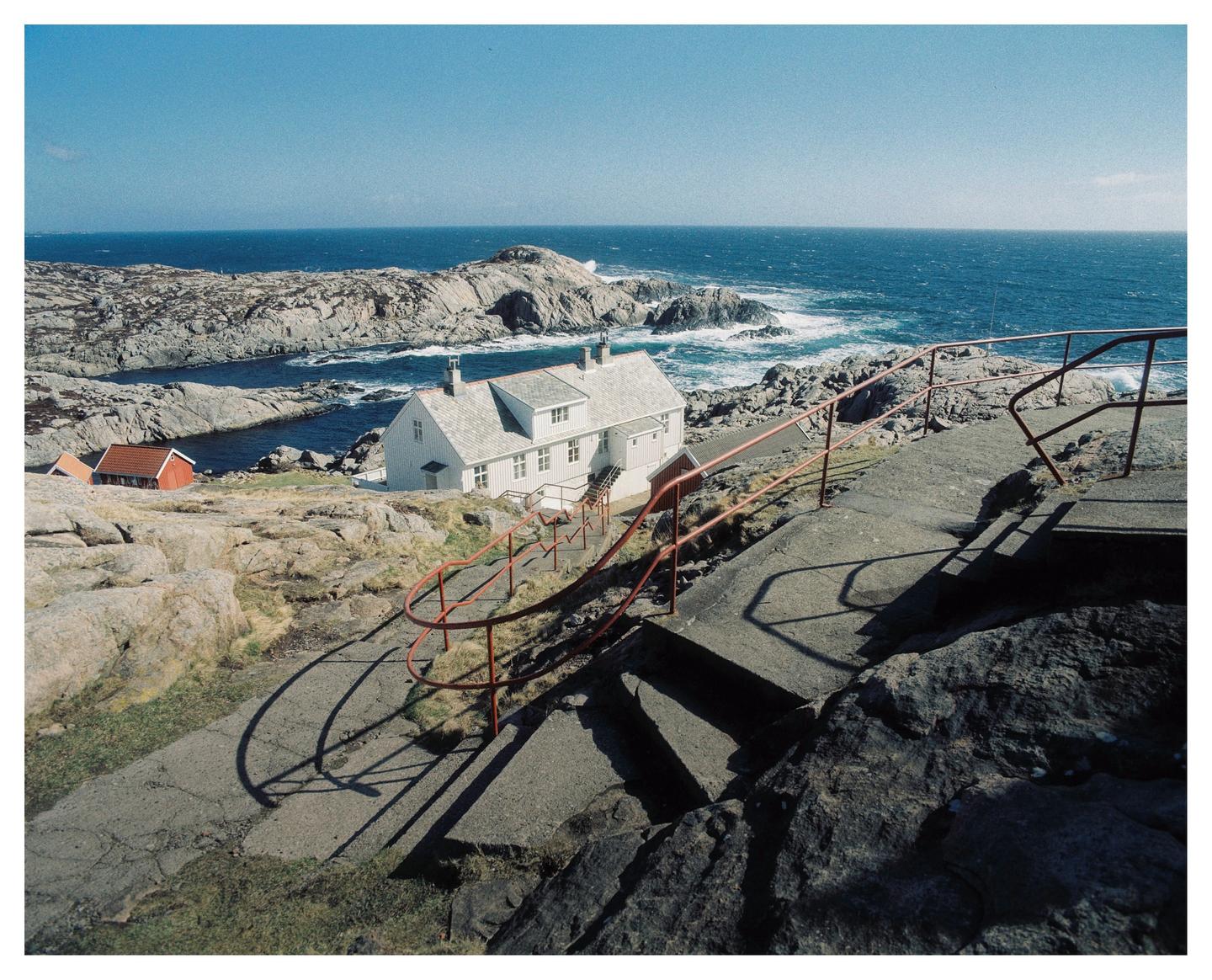 A white house with a red roof sits on a rocky coastline overlooking the ocean, with a concrete path and red railings in the foreground.