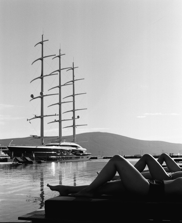 Two people relax by a pool with a large sailboat in the background.