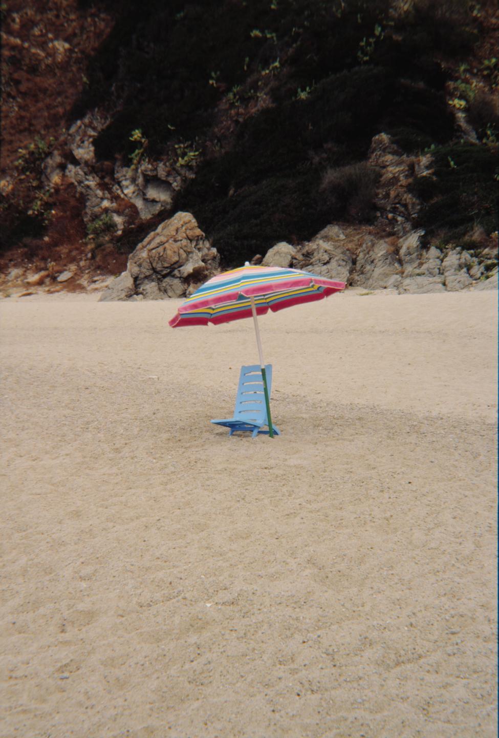 A colorful striped beach umbrella and a blue beach chair sit on a sandy beach with a rocky hill in the background.