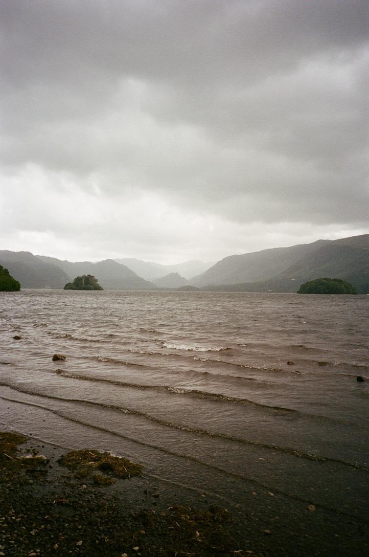 A lake with small waves and two islands is surrounded by mountains under a cloudy sky.