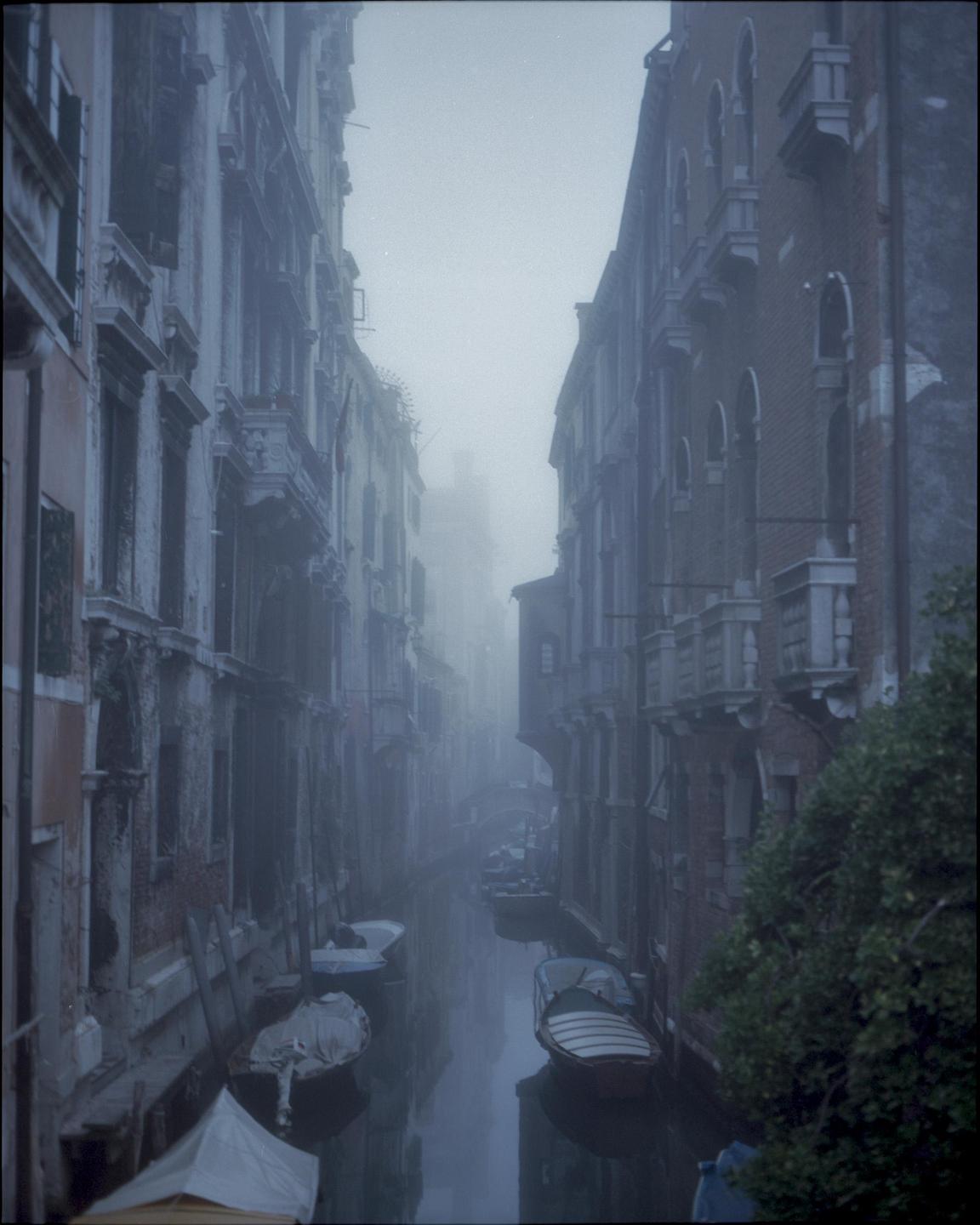 A narrow canal with boats is visible between tall buildings on a hazy day in Venice.