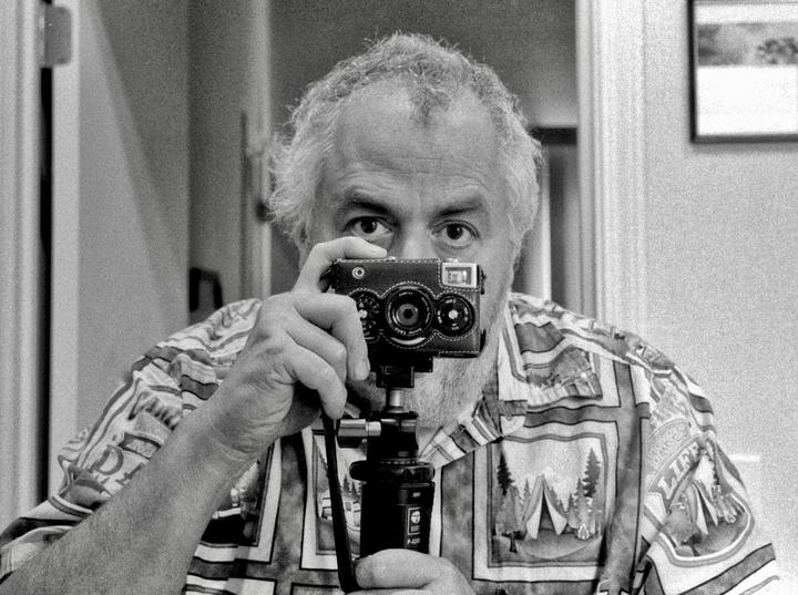 A man with a beard and patterned shirt holds a camera on a tripod in front of his face, taking a self-portrait in a mirror.