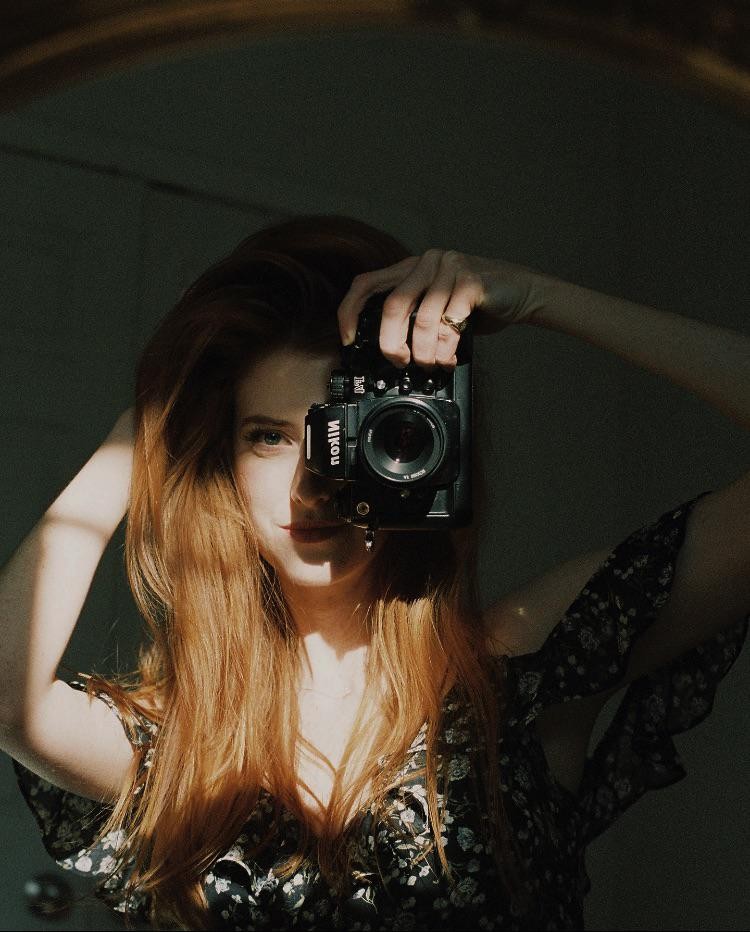 A woman with long red hair holds a camera in front of her face, obscuring one eye, in a self-portrait.