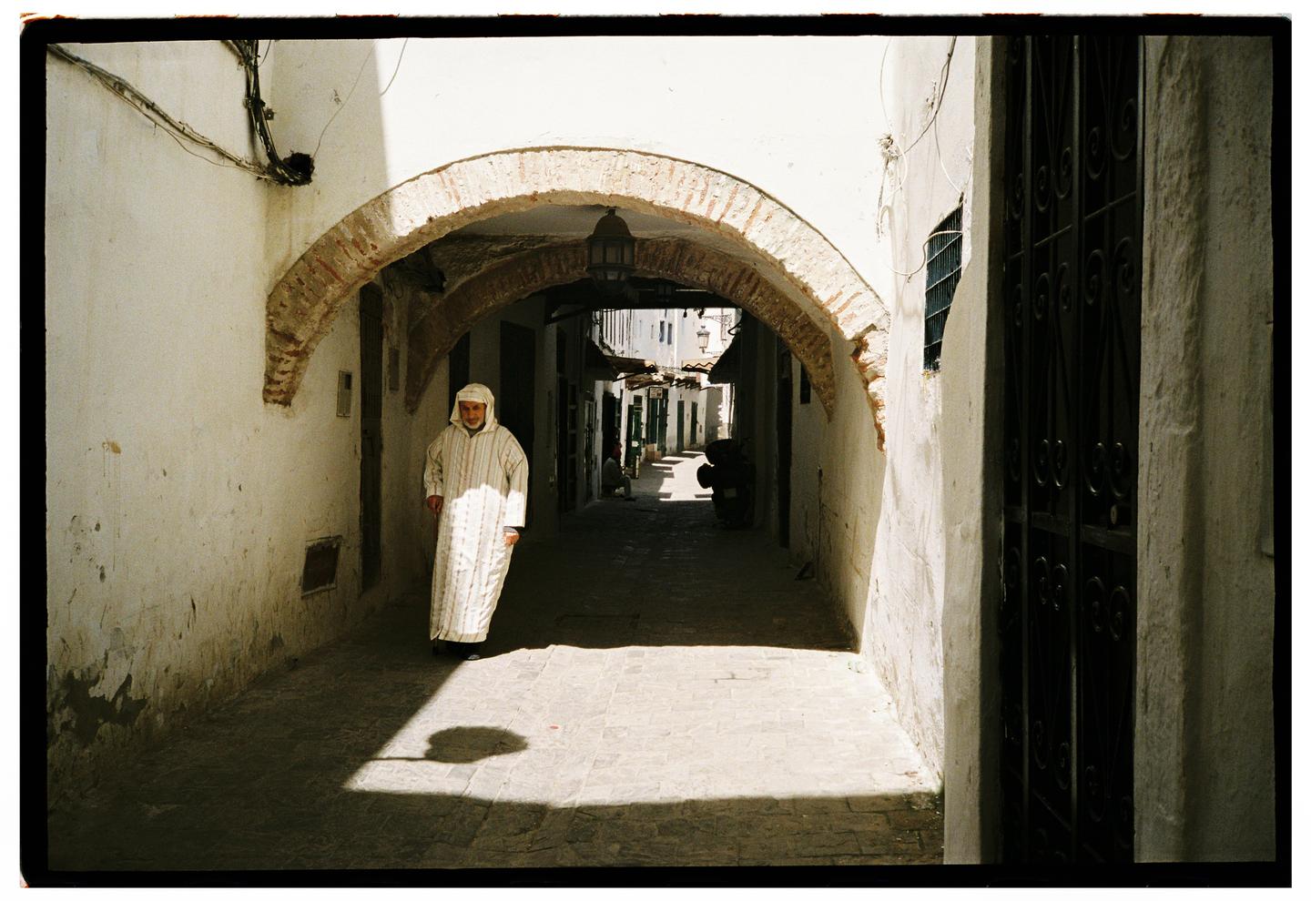 A man in a traditional robe walks through a sunlit arched alleyway in a medina.