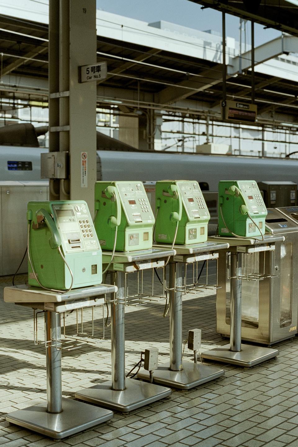 Four green payphones stand in a row on a train platform with a train in the background.